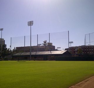 A sunlit baseball field with large industrial netting, surrounded by bleachers and buildings, under a clear blue sky.