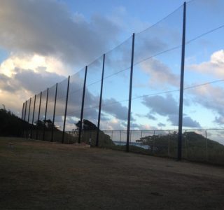 A row of tall poles with industrial netting at a coastal baseball field under a cloudy sky at twilight.