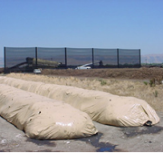 Large beige sandbags in foreground with a tall, golf netting fence behind them under a clear blue sky.
