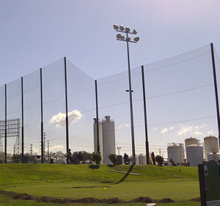 A golf driving range with high golf netting and lighting poles, set against a backdrop of industrial tanks and a clear sky.