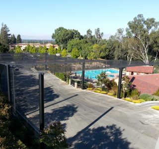 A view of a shaded walkway overlooking a swimming pool and landscaped area, surrounded by trees and buildings under a clear sky, complemented by aesthetically placed landfill netting.