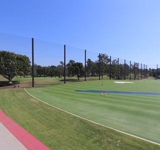 An outdoor sports field with a large green artificial turf area surrounded by sports netting and trees under a clear sky.