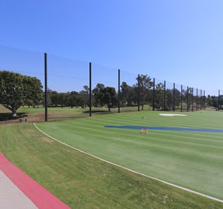 A wide view of a sports field with synthetic turf surrounded by tall sports netting, trees in the background, under a clear blue sky.