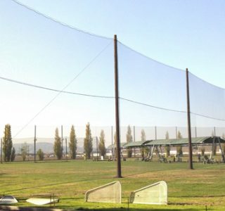 Baseball field with high protective nets, picnic areas, and trees in the background under a clear sky, surrounded by industrial netting.