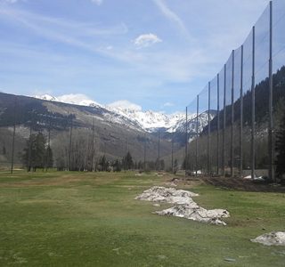A golf driving range with grassy grounds bordered by tall sports netting, with mountains and patches of snow in the background.